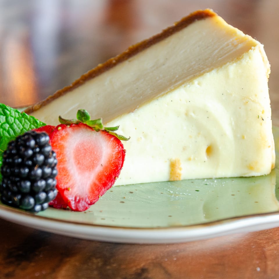 Slice of creamy cheesecake on a plate, garnished with fresh strawberries, blackberry, and mint leaves.