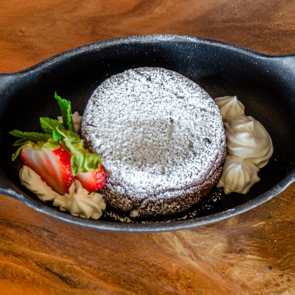 Chocolate dessert dusted with powdered sugar, served with whipped cream, strawberries, and mint in a black dish.