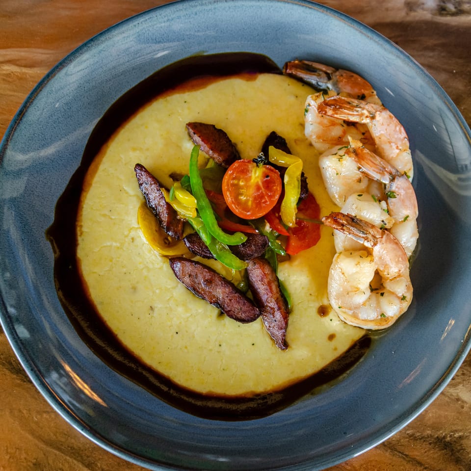 Creamy polenta topped with grilled shrimp, sliced sausage, colorful peppers, tomatoes, and dark sauce on a blue plate.