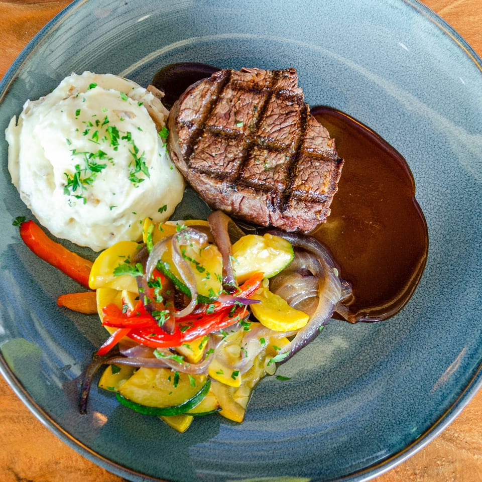 Grilled steak with mashed potatoes, sautéed vegetables, and brown sauce on a blue plate.
