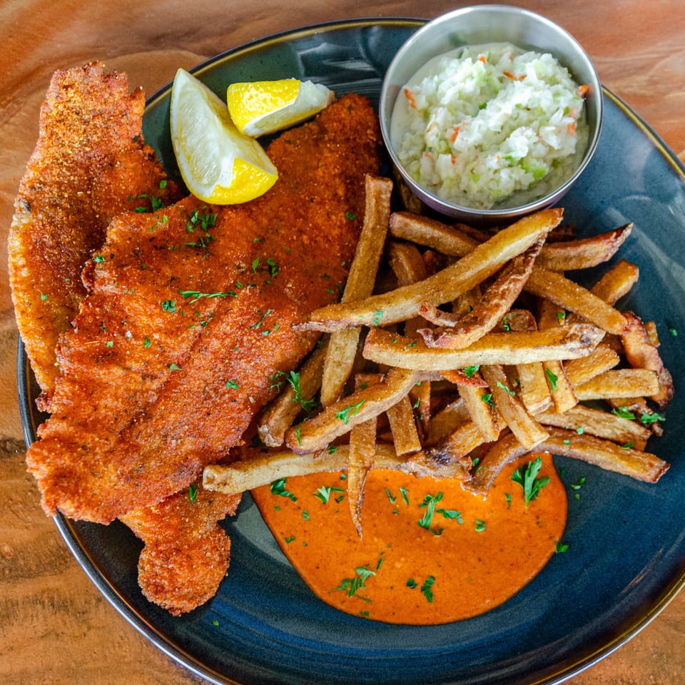 Crispy breaded fish fillets with fries, lemon wedges, coleslaw, and dipping sauce on a black plate.
