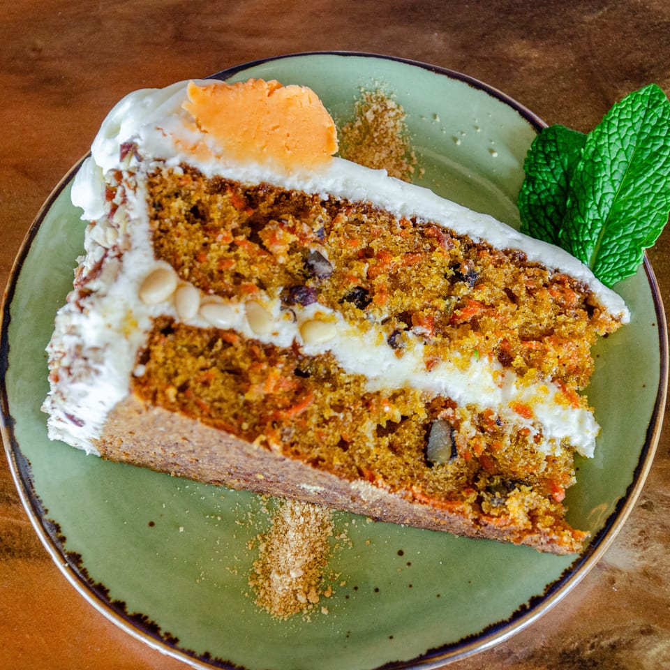 Slice of layered carrot cake with cream frosting, crumb topping, orange garnish, and fresh mint on a green plate.