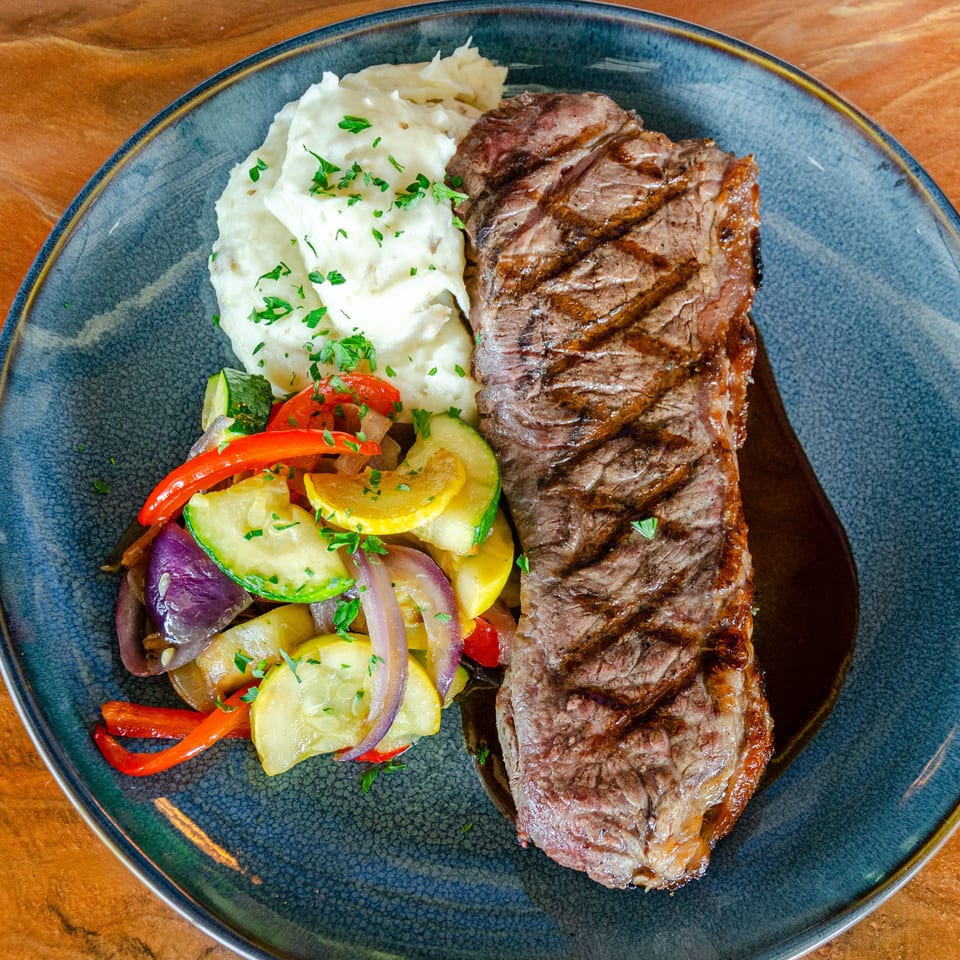 Grilled steak with mashed potatoes and colorful sautéed vegetables on a blue plate atop a wooden table.