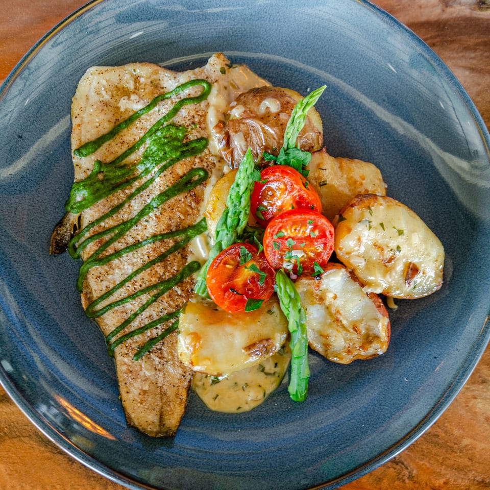 Seared fish fillet with green sauce, roasted potatoes, asparagus, and sliced tomatoes on a blue plate.