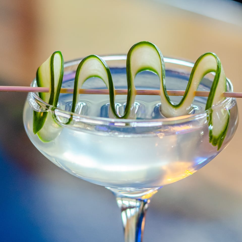 Clear cocktail in a stemmed glass garnished with curled cucumber slices and a pink straw.